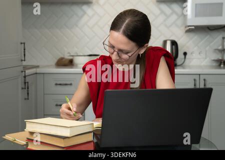 Focused female freelancer wearing glasses and a red shirt working from her home kitchen, using a laptop and taking notes in a notebook, with several b Stock Photo