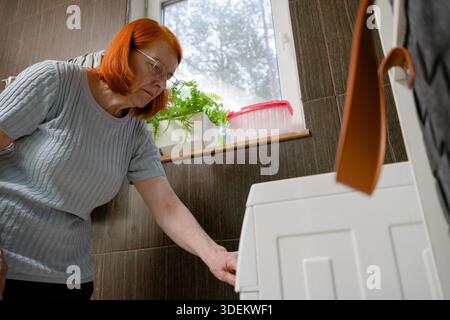 A low-angle shot from below showing a senior female pressing the start button on a front-load washing machine in a domestic setting. Stock Photo