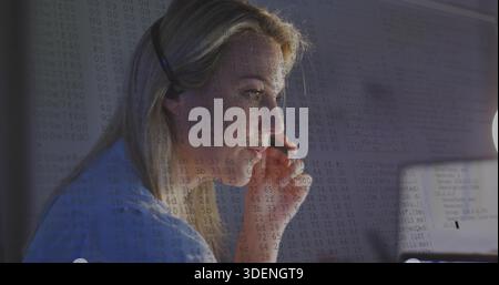 Leaning woman in light-blue top adjusting headset and studying laptop at home office, code overlay Stock Photo