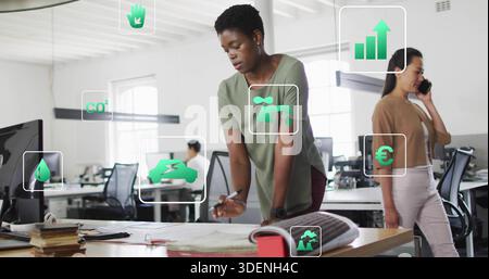 Leaning woman in olive-green top reviewing plans and making notes in open-plan office with AR icons Stock Photo