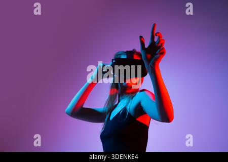Woman navigating virtual reality space with VR headset and expressive hand gestures in futuristic purple and red lighting Stock Photo
