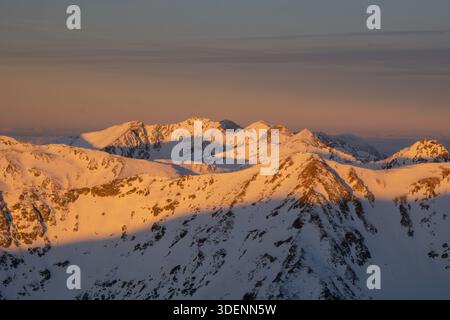 Aerial view of snow-capped peaks kissed by the warm glow of the setting sun, contrasting against the cool, shadowy valleys below, Nizna Bystra, Zilina Stock Photo