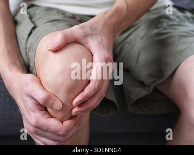 A middle-aged man sitting on a couch, holding his knee and leg with both hands in pain. Close up. Stock Photo