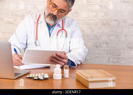 Doctor wearing a white lab coat using a digital tablet and writing, with pills on desk Stock Photo