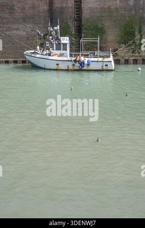A small, white fishing boat with a blue bottom line, moored on a high, algae-covered quay wall in the harbor, while some seagulls swim on the calm, br Stock Photo