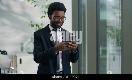 Smiling African American businessman using smartphone in office business man ethnic male guy looking at mobile phone cheerful professional Stock Photo