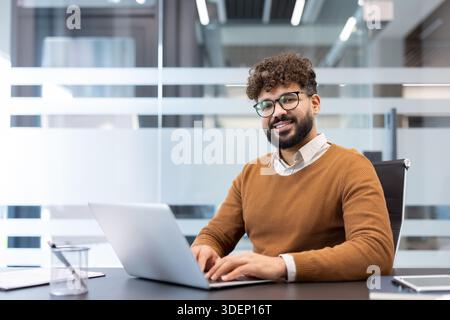 Young man with beard and glasses smiling at camera, sitting at a desk and using a laptop in a contemporary office environment, embodying professionalism and success Stock Photo