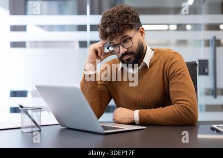 Young bearded businessman in glasses concentrating on his laptop at a modern office desk, thoughtfully analyzing data and planning solutions for a corporate project or strategy Stock Photo
