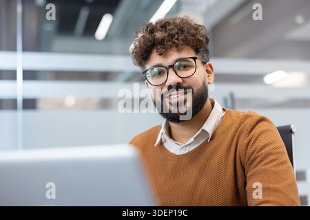 Confident young man with beard and glasses smiling at the camera, sitting at a desk with a laptop, representing business success and professional achievement Stock Photo