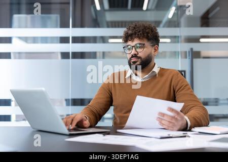 Young businessman analyzing data and typing on a laptop, managing paperwork and responsibilities while staying productive in a modern office environment Stock Photo