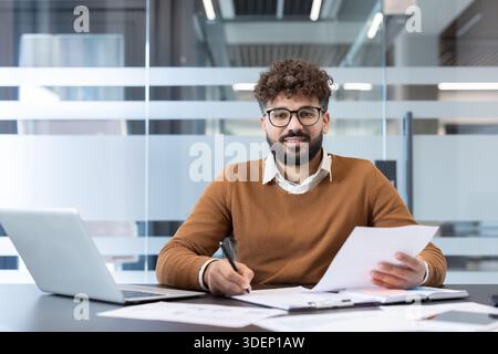 Young businessman with curly hair and glasses smiling at the camera while writing notes and holding paperwork, working at a modern office desk with a laptop Stock Photo