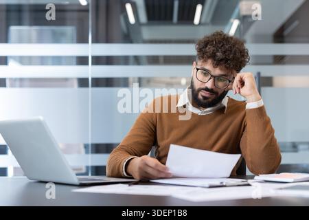 Young businessman with curly hair and glasses sitting at a desk, reviewing documents and planning while working on a laptop in a contemporary office environment Stock Photo