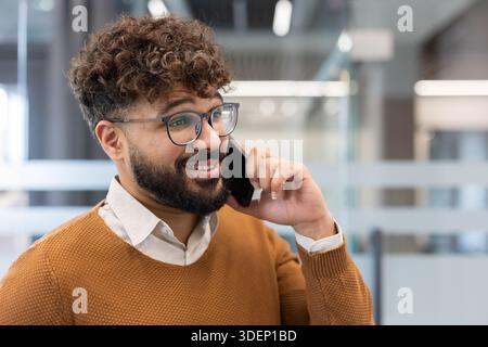 Young professional man with curly hair, eyeglasses and beard smiling while talking on a smartphone in a modern office, confident and approachable during a work call Stock Photo