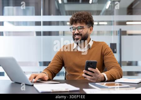Smiling young bearded man wearing glasses sitting at a desk, looking at his smartphone while simultaneously typing on a laptop, working efficiently in a modern office environment Stock Photo