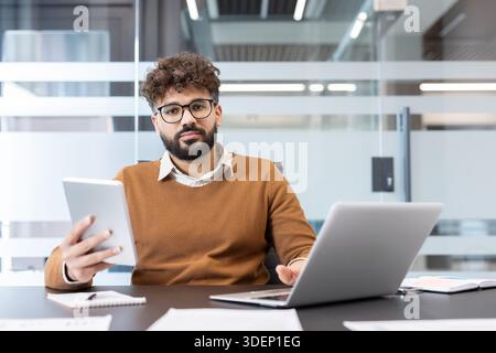 Young professional with glasses and curly hair working on laptop and tablet at a modern office desk, focused on digital tools and productive startup-style business planning Stock Photo
