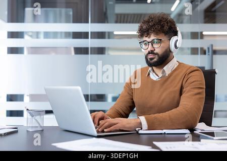 Young indian man in a brown sweater and glasses sitting at a desk, typing on a laptop and wearing white headphones, concentrating on his work in a contemporary office environment Stock Photo