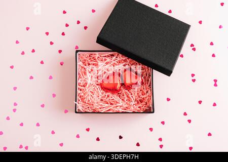 Open black gift box with red heart chocolates inside on pink background with scattered confetti. Valentines day concept. Top view, flat lay. Stock Photo