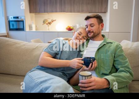Smiling relaxed couple of man and woman embracing each other and resting together on couch at home. Stock Photo
