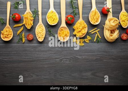 Different types of raw pasta in spoons, tomatoes, rosemary and thyme on wooden table, flat lay. Space for text Stock Photo