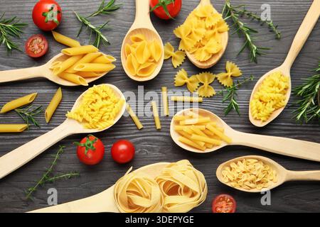 Different types of raw pasta in spoons, tomatoes, rosemary and thyme on wooden table, flat lay Stock Photo