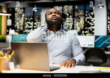 Black male professional does self massage to relieve neck stiffness and discomfort, dealing with tension due to incorrect office posture after hours. Stretching his upper back muscles. Stock Photo