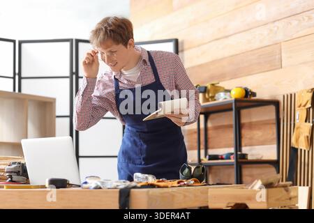 Young carpenter with notebook using laptop at table in workshop Stock Photo