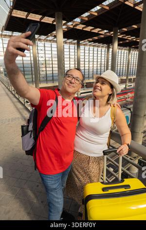 Smiling adult travel couple taking a selfie and looking at a smartphone while standing inside a modern train station with luggage. Selfie travel momen Stock Photo