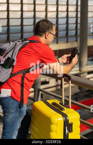 Mature male traveler using a smartphone at a train station while standing next to a yellow suitcase. Concept of travel, technology, urban mobility and Stock Photo