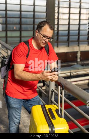Adult man standing at an urban train station, holding a yellow suitcase and looking at the trains below while checking his phone during a travel momen Stock Photo