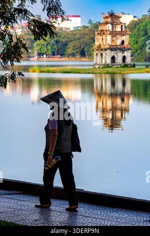 Vietnamese lady wearing bamboo hat walking past Turtle Tower, Hoan Kiem lake, Hanoi, Vietnam Stock Photo