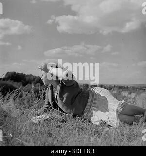 Slaviansk, Donetsk region, Ukraine, USSR. 1950s. Artistic archival photo of Valentina Timofeevna Kravelis resting on the high bank of the Kazeny Torets river. The young woman lies on the grass in a stylish striped dress, shielding her eyes from the bright summer sun with her hand. The vast landscape of the river valley opens up in the background. The image captures the aesthetics of Soviet fashion, the carefree mood of youth and the serene atmosphere of the peaceful Donbas during the stable period of life before the war. Stock Photo