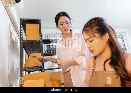 Two young women are working together in a small business office, surrounded by shelves of packages and documents. They discuss tasks, check inventory, Stock Photo