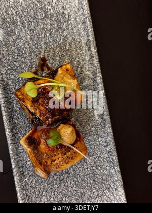 Two seared gourmet cubes, possibly tofu or fish, garnished with microgreens and sauce, served on a gray stone plate. Stock Photo