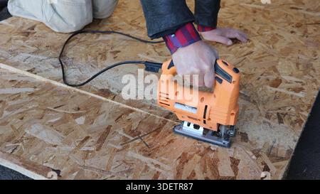 A partially visible white man kneeling with an orange electric jigsaw in his right hand and sawing a sheet of OSB plywood lying on the ground Stock Photo