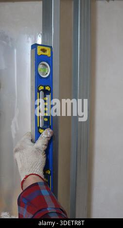 A gloved hand vertically holds a blue level on a metal profile in a room structure and measures the angle of inclination, taking measurements Stock Photo
