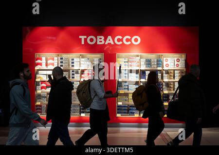 People walk past a tobacco display at an airport duty-free shop, likely in Europe, showcasing various cigarette brands. Stock Photo