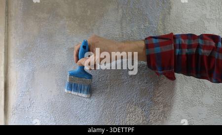 Hand of a man in a plaid shirt holds a blue brush and applies liquid primer on a textured plastered gray wall, close-up, priming a home wall Stock Photo