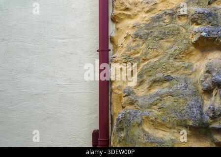 A red drainpipe runs along the junction of two contrasting wall surfaces, a smooth beige plaster and a rough yellow stone texture, in Roth, Germany. The composition emphasizes material contrast and color geometry. Stock Photo