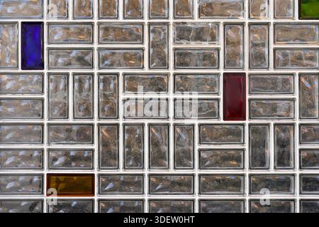 A section of a patterned glass block wall with rectangular translucent elements in Roth, Germany. Among the mostly grey-tinted glass blocks, a few are colored in red, blue, green, and amber, creating subtle visual accents within the geometric grid. Stock Photo