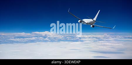 White passenger airplane flying through clear blue sky above a layer of clouds Stock Photo
