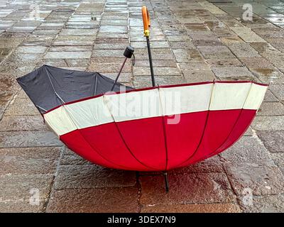 Venice, Veneto, Italy. Two abandoned umbrellas lie on the pavement. The red and white umbrella is open. The ground is wet from rain. Stock Photo