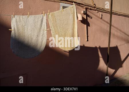 Burano, Venice, Veneto, Italy. Two rugs hang on a clothesline outside. The light brown wall casts a long sharp shadow. Stock Photo
