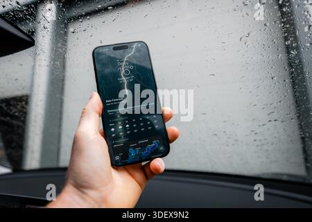 A hand holds a smartphone displaying a severe weather forecast inside a car, with raindrops on the windscreen, conveying stormy conditions, travel dis Stock Photo