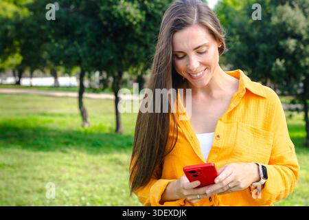 Smiling woman in yellow shirt holding smartphone and looking at camera outdoors Stock Photo