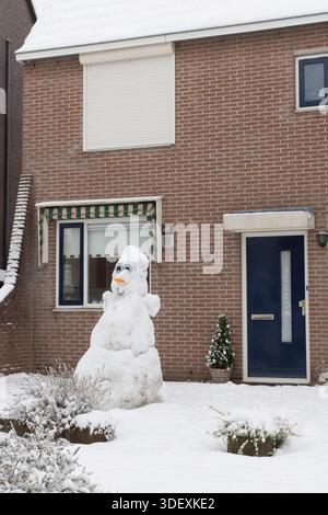 A cheerful snowman stands in the snowy front yard of a brick house in the Netherlands, beside a striped awning window and a blue door, creating festiv Stock Photo
