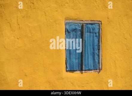 A blue window made of corrugated iron, painted blue, and set into a yellow painted wall. Andranovory, Madagascar.  The contrasting colours are typical of rural Malagasy villages, where residents often use inexpensive, available materials like corrugated metal and bright paints to build and decorate their homes, adding charm and character to the local architecture. Stock Photo