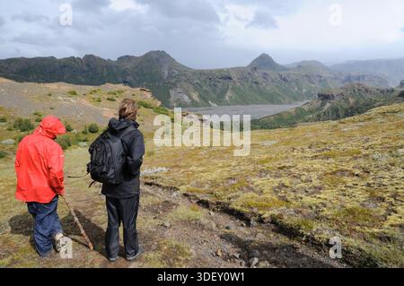 Two young people standing on a hiking trail overlooking a rugged mountain landscape in Iceland, wearing outdoor clothing in changeable weather. Stock Photo