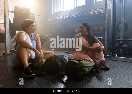 Diverse female friends sitting on rubber mat resting between sets near kettlebells and gym bags Stock Photo