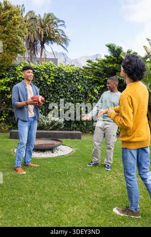 Diverse male friends standing in backyard holding football near fire pit in sneakers, copy space Stock Photo