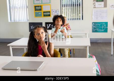 Diverse children classmates sitting at desks in classroom, pondering with silver laptop nearby Stock Photo